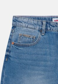 Modri traper hlače z klasičnim pet-pocket dizajnom, vidnimi šivi in kovinsko etiketo blagovne znamke na sprednjem žepu.