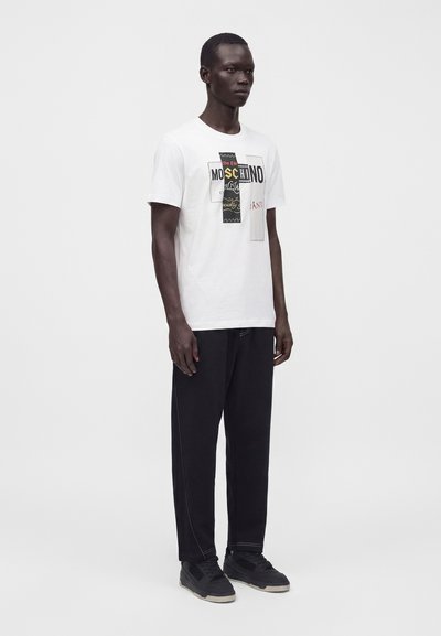 Άνδρας στέκεται φορώντας ένα λευκό graphic t-shirt Moschino, μαύρο παντελόνι με ορατές ραφές και μαύρα αθλητικά παπούτσια σε απλό λευκό φόντο.