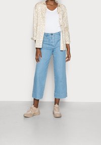 Světle modré zkrácené džíny s vysokým pasem, v kombinaci s béžovou košilí s vzorem a béžovými teniskami. Oblečení má volný, uvolněný střih.