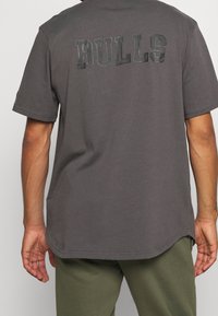 Muž v tmavě šedé košili s krátkým rukávem s nápisem „BULLS“ na horní části zad a v olivově zelených kalhotách, stojící s uvolněnýma rukama.