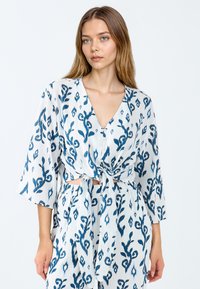 Weiße Bluse mit blauem, floralem Muster, V-Ausschnitt, gebundenem Front und lockeren Ärmeln. Leichter, strukturierter Stoff fällt sanft.