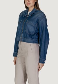 Giacca di jeans tagliata di colore azzurro chiaro, con due tasche frontali e una zip centrale. Abbinata a pantaloni larghi di colore chiaro.