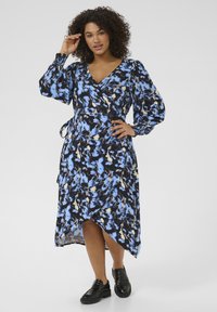 Robe portefeuille en bleu, noir et motifs floraux crème ; dotée de manches bouffantes longues, d'une taille cintrée et d'un ourlet asymétrique. Portée avec des chaussures noires.