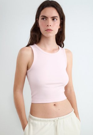 Jonge vrouw met lang donker haar draagt een lichtroze mouwloze crop top en lichtgekleurde broek, staand met handen in de zakken tegen een effen achtergrond.