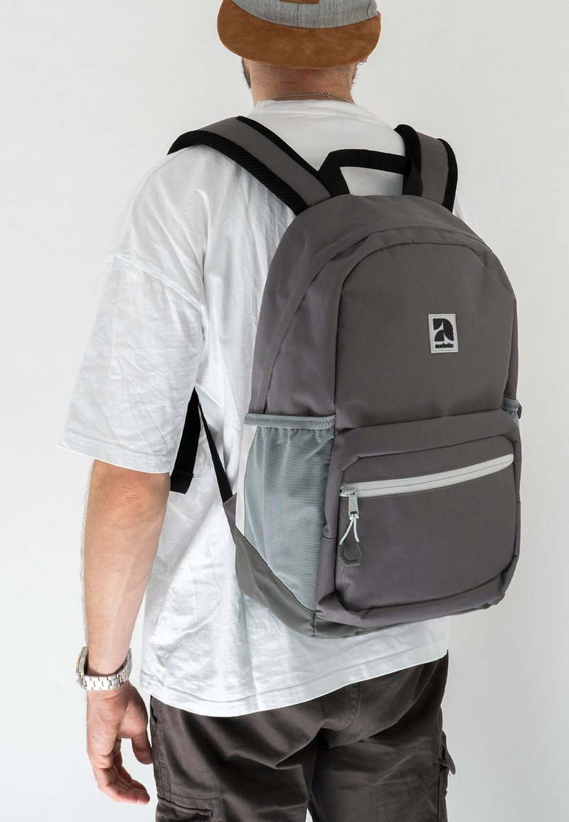 Gray fabric backpack with a main compartment and front pocket. Features mesh side pocket and black adjustable straps. Simple design with logo.