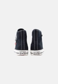 Μαύρα ψηλά αθλητικά παπούτσια από καμβά με λευκή ραφή και λαστιχένιες σόλες. Διαθέτουν ένα λογότυπο σε πατς και ένα διακοσμητικό γούρι στο πλάι.