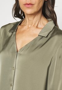 Blouse vert olive satinée avec col cranté, fermeture à boutons sur le devant et un éclat doux. Présente une coupe décontractée et une texture lisse.