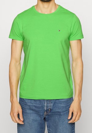 Grünes kurzärmliges T-Shirt mit rundem Ausschnitt und kleinem Logo auf der Brust. Hergestellt aus leichtem, glattem Stoff. Kombiniert mit blauen Jeans.