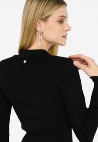 Giacchetto nero a coste con collo a giacca e maniche strutturate. Orecchini a cerchio in argento molto evidenti e un piccolo dettaglio metallico sul retro.