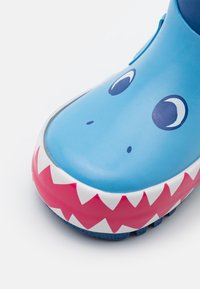Blauwe rubberen schoen met een ronde neus, voorzien van een haaienontwerp, een patroon van witte tanden, roze accenten en grote cartoonogen.