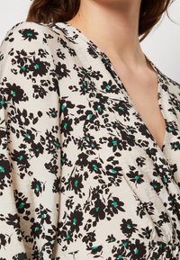 Top enroulé floral en tissu léger; présente un motif floral noir et vert sur un fond crème, avec un décolleté en V profond et un détail froncé.