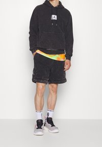 Felpa nera con cappuccio e tasca frontale, abbinata a pantaloni corti neri. Sottogiacca in rete dai colori vivaci e scarpe da ginnastica grigie.
