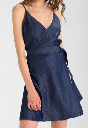 Mujer con un vestido cruzado sin mangas de mezclilla azul oscuro con tirantes finos y un cinturón a juego atado a la cintura.