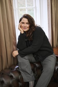 Pikkade tumedate juustega naine mustas sviitris ja hallides pükstes istub pruunil nahkdiivanil akna ääres, mille ees on beežid kardinad, mõtlikult naeratades.