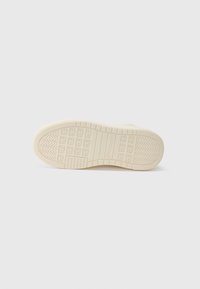 Semelle de sneaker en caoutchouc crème avec une surface texturée, présentant des motifs circulaires et en zigzag pour l'adhérence, ainsi qu'un logo de marque en relief.