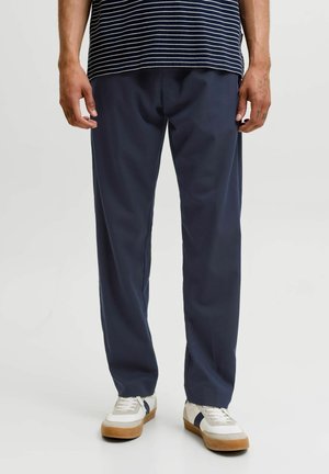 Mand iført marineblå bukser med lige ben, hvide sneakers med gummisåler og en navyblå og hvidstribet skjorte, stående på en ensfarvet baggrund.