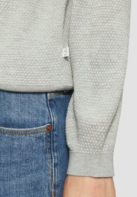 Hellegrüner strukturierter Pullover mit einem Wabenmuster, der eng anliegende Ärmel und ein kleines Etikett hat, kombiniert mit blauen Denim-Jeans.