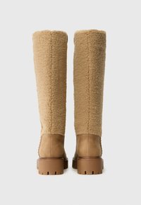 Botas hasta la rodilla en beige con parte superior de suave y texturizada piel de oveja y sección inferior de piel lisa, con suela de goma gruesa y texturizada.