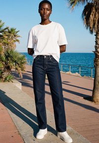 Femme portant un t-shirt blanc, un jean bleu foncé et des baskets blanches, debout près des palmiers au bord de l'océan par une journée ensoleillée.