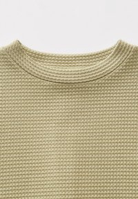 Tissu en tricot gaufré beige clair avec un col rond, montrant les détails de la texture et des coutures sur une surface plane.