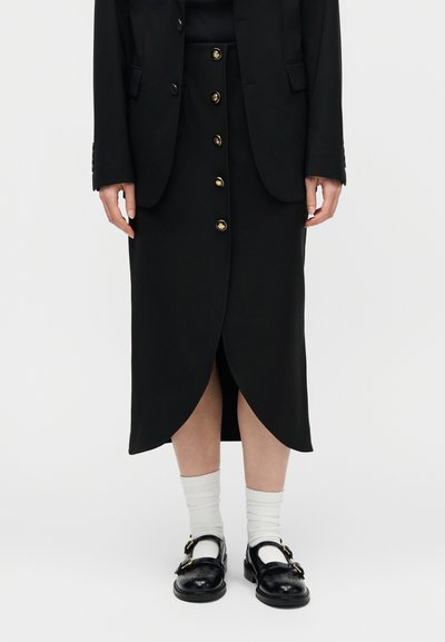 Μαύρη midi φούστα με χρυσά κουμπιά στο μπροστινό μέρος, τελείωμα σε σχήμα τουλίπας, συνδυασμένη με μαύρα παπούτσια με αγκράφες και άσπρες κάλτσες, φοράει μαύρο σακάκι.