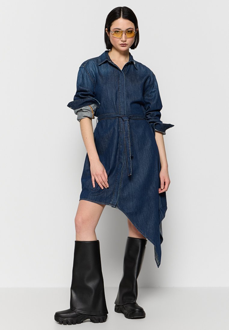 Robe en denim avec taille ceinturée, ourlet asymétrique et manches retroussées. Associée à des bottes en caoutchouc noires jusqu'aux genoux.