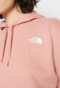 Personne portant un sweat à capuche rose poudré avec cordons, avec le logo The North Face en blanc sur le haut de la poitrine.