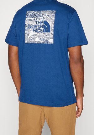 Osoba nosící modré tričko s bílým abstraktním potiskem hory a slunce a logem "The North Face" na zádech, doplněná hnědými kalhotami.