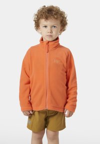 Chaqueta de forro polar naranja con un cuello alto y cremallera frontal, que presenta el logo en el pecho, combinada con pantalones cortos caqui. Textura suave en toda la prenda.