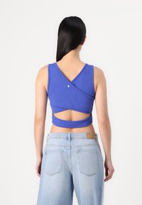 Blaues ärmelloses Crop-Top mit einem gekreuzten Rückendesign und einer zentralen Knopfdetail, kombiniert mit hellblauen hochgeschnittenen Jeans.