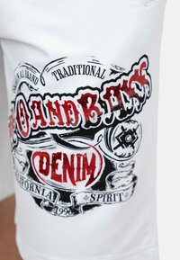 Witte t-shirt met een grafisch ontwerp in rood en zwart, met de tekst "Originele Merk Traditionele Denim," vergezeld van decoratieve accenten en patronen.