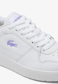 Białe skórzane sneakersy o gładkiej fakturze, z fioletowym logo Lacoste i białymi sznurowadłami. Okrągły czubek i gumowa podeszwa.