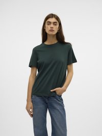 Ung kvinne med langt brunt hår iført en mørkegrønn kortermet skjorte og blå jeans, stående med én hånd i lommen.