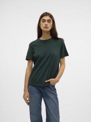 Ung kvinne med langt brunt hår iført en mørkegrønn kortermet skjorte og blå jeans, stående med én hånd i lommen.