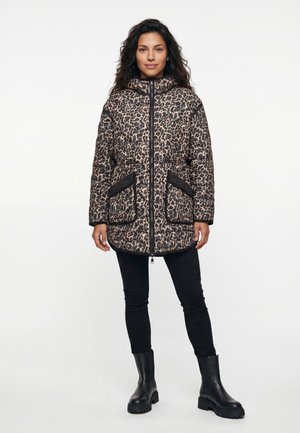 Frau steht und trägt eine lange gesteppte Jacke mit Leopardenmuster, schwarze Hose und schwarze klobige Stiefel vor schlichtem Hintergrund.