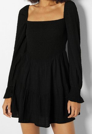 Femme portant une robe noire à manches longues avec un décolleté carré et un corsage froncé, debout devant un fond blanc uni.