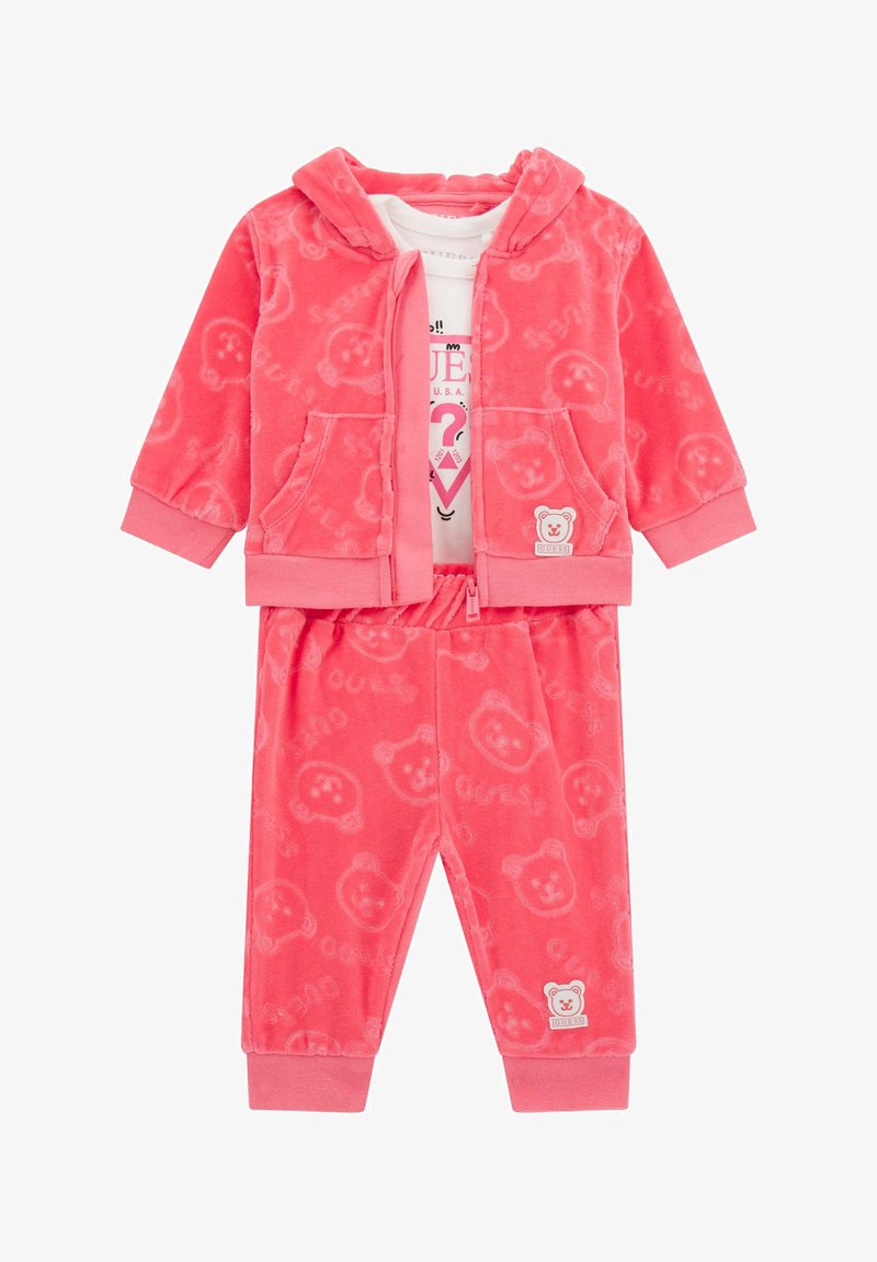 Ružový, plyšový dvojdielny outfit pozostávajúci z mikiny na zips so vzorom medveďa a zodpovedajúcich nohavíc, obidva vybavené elastickými manžetami a pásom.