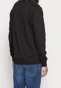 Sudadera negra con cuello redondo, tela texturizada y mangas largas. Combinada con unos vaqueros de mezclilla azul que tienen dobles bolsillos traseros.