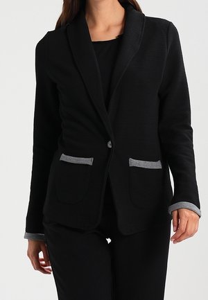 Femme portant un blazer noir avec un bord gris sur les poches et les poignets, associé à un pantalon noir, sur un fond uni.