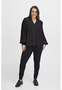Černá blůza s dlouhým rukávem a véčkovým výstřihem, sklady na textuře a knoflíkovým zapínáním. V kombinaci s černými slim-fit kalhotami a černými botami.
