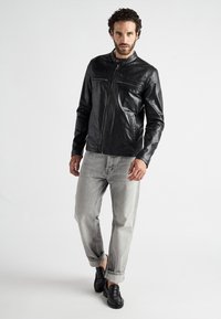 Schwarze Lederjacke mit Stehkragen und Reißverschlusstaschen, kombiniert mit hellgrauen Jeans und schwarzen Slippern.