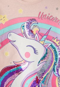 Jasnoróżowa koszulka z grafiką jednorożca, z cekinową grzywą w żywych kolorach, otoczona akcentami tęczy i gwiazdek.