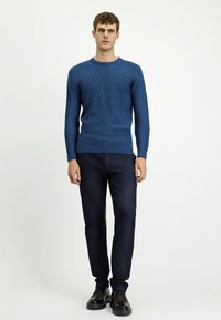 Niebieski dzianinowy sweter z fakturowanym wzorem, okrągłym dekoltem i ściągaczami przy mankietach, zestawiony z ciemnymi jeansami i czarnymi butami. Gładkie tło.