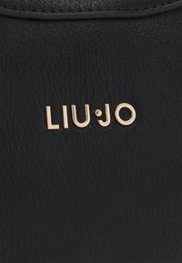Surface en cuir noir texturé avec des lettres métalliques dorées du logo de la marque "LIU·JO" centrées près du bord supérieur.