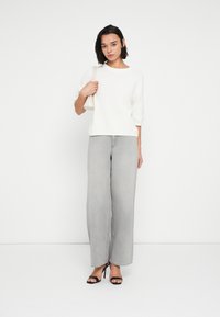 Άσπρο πλεκτό πουλόβερ με κοντά μανίκια, σε συνδυασμό με ανοιχτό γκρι τζιν φαρδιάς γραμμής. Το μοντέλο κρατά μια μικρή άσπρη τσάντα και φορά μαύρα σανδάλια με ψηλό τακούνι.