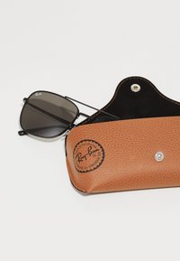 Ochelari de soare Ray-Ban cu rame negre și lentile închise, lângă o husă din piele maro texturată, cu un logo embosat și închidere cu clichet argintie.