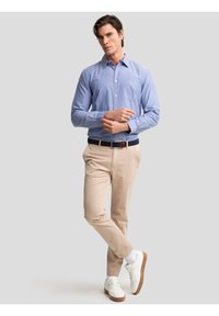 Bărbat purtând cămașă bleu deschis cu nasturi, pantaloni bej, adidași albi și curea bleumarin, stând cu picioarele încrucișate pe un fundal simplu.