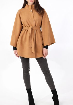 Frau trägt einen kamelfarbenen Mantel mit Gürtel und weiten Ärmeln, dunkelgraue Skinny Jeans und schwarze Stiefeletten, steht vor weißem Hintergrund.