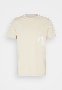 Camiseta de algodón color crema con cuello redondo y mangas cortas. Presenta un sutil logo blanco "ck" en la parte izquierda del pecho. Diseño sencillo.