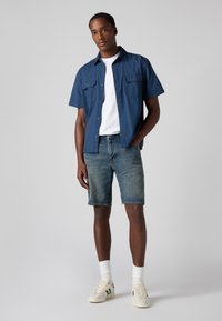 Cămașă cu mânecă scurtă albastră închis, tricou alb dedesubt, pantaloni scurți din denim și adidași crem cu accente verzi. O ținută simplă și lejeră.